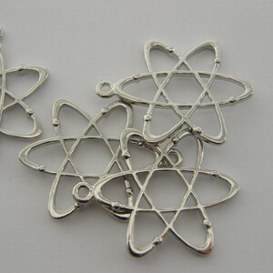 Op de afbeelding: Zilverkleurige atoomvormige bedels. Elke bedel heeft een centrale stervormige structuur met draaiende ringen en kleine kraalachtige accenten. De bedels hebben een lus aan de bovenkant om aan sieraden of andere knutselwerken te bevestigen.