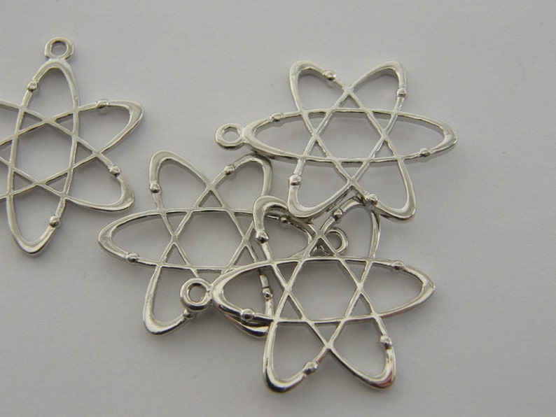 Op de afbeelding: Zilverkleurige atoomvormige bedels. Elke bedel heeft een centrale stervormige structuur met draaiende ringen en kleine kraalachtige accenten. De bedels hebben een lus aan de bovenkant om aan sieraden of andere knutselwerken te bevestigen.