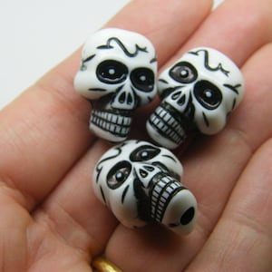 May include: Three white plastic skull beads with black painted details. The skulls have black eyes, a black nose, and a black mouth.