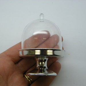 May include: A small, clear plastic cake dome with a silver base. The dome is on a pedestal with a round base.