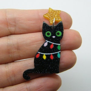 Puede incluir: Adorno navideño de gato negro con purpurina con una estrella dorada con purpurina en la parte superior. El gato está decorado con luces de colores.