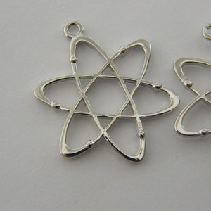 Op de afbeelding: Close-up van verschillende zilverkleurige atoombedelhangers. Elke hanger heeft een stervormig ontwerp met een centrale zeshoek en draaiende ovale vormen, geaccentueerd met kleine baldetails. De hangers hebben een lus aan de bovenkant om te bevestigen.