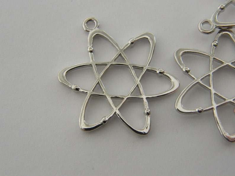 Op de afbeelding: Close-up van verschillende zilverkleurige atoombedelhangers. Elke hanger heeft een stervormig ontwerp met een centrale zeshoek en draaiende ovale vormen, geaccentueerd met kleine baldetails. De hangers hebben een lus aan de bovenkant om te bevestigen.