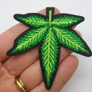 May include: Embroidered patch in the shape of a cannabis leaf. The leaf has a detailed design with varying shades of green and a black border. This patch is likely for decorative purposes, such as adding to clothing or accessories.