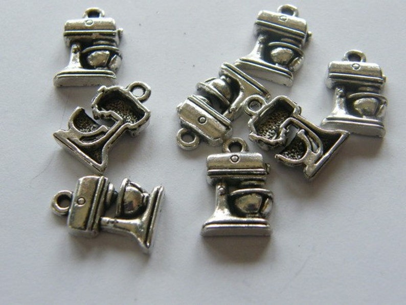 May include: A group of eight silver-toned metal charms shaped like stand mixers. The charms have a loop at the top for attaching to a necklace, bracelet, or other jewelry.