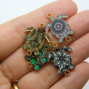 May include: Four gold-tone metal turtle charms with different patterned designs. The turtles have a black and white mandala design, a green and black spotted design, a gold and green spotted design, and a pink, green, and white mandala design.