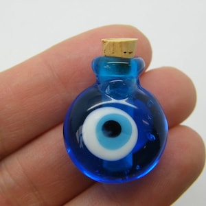 Puede incluir: Una pequeña botella de vidrio azul con un tapón de corcho y un amuleto de ojo turco blanco en su interior. La botella es redonda y tiene una superficie lisa.