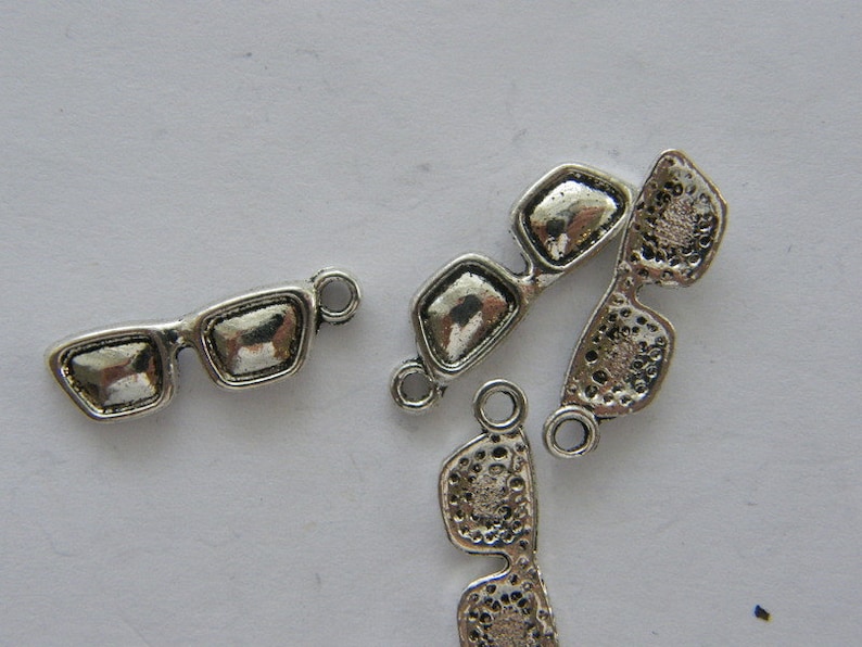 May include: Silver-toned metal charms shaped like sunglasses. The charms have a loop at the top for attaching to jewelry or other crafts.