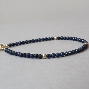 Peut inclure: Un bracelet délicat composé de petites perles bleu foncé facettées, entrecoupées de perles dorées. Le bracelet est doté d'un fermoir doré et d'un petit charm. Le bijou est présenté sur un fond gris neutre.