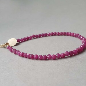 Op de afbeelding: Een delicate armband met kleine, gefacetteerde, dieproze edelstenen die aan elkaar zijn geregen. De armband heeft een goudkleurige sluiting en een kleine, ronde bedel. De sieraden worden gepresenteerd op een neutrale grijze achtergrond.
