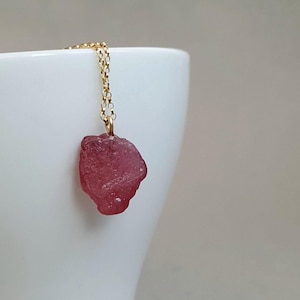 Puede incluir: Un collar de cadena dorada con un colgante de piedra preciosa rosa en bruto. El colgante tiene una forma rugosa e irregular, y la cadena es delicada. El collar se muestra sobre un fondo blanco.