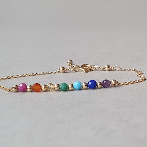 Puede incluir: Un brazalete de cadena dorada con siete cuentas de piedras preciosas de colores. Las cuentas son rojas, naranjas, amarillas, verdes, azules, turquesas y moradas.