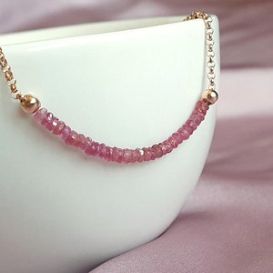 May include: A delicate necklace with a strand of small, pink gemstones. The gemstones are strung in a curved line, with a small gold bead at each end. The chain is a delicate gold colour. The necklace is displayed against a white background.