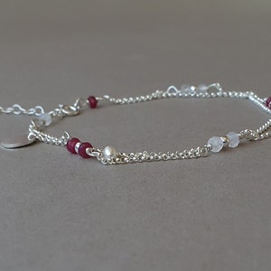 Op de afbeelding: Een delicate zilveren enkelketting met een klein zilveren bedeltje, met rode en witte kralen.