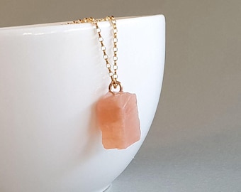 Può includere: Una collana con catena color oro con un ciondolo di pietra preziosa rosa chiaro grezza. Il ciondolo ha una forma approssimativamente rettangolare con una superficie strutturata. La collana è esposta su sfondo bianco.