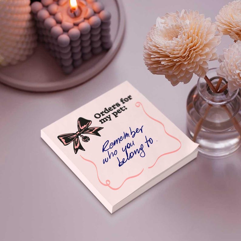 Peut inclure: Bloc-notes carr&eacute; avec le texte "Orders for my pet: Remember who you belong to." Un n&oelig;ud noir et rose est au-dessus du texte. Le bloc-notes est sur une surface violet clair, avec une bougie et des fleurs en arri&egrave;re-plan.