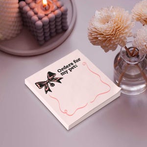 Peut inclure: Un bloc-notes carr&eacute; rose clair avec le texte "Orders for my pet:" et une illustration d'un n&oelig;ud noir. Le bloc-notes a une bordure rose ondul&eacute;e. Des fleurs d&eacute;coratives et une bougie sont en arri&egrave;re-plan.