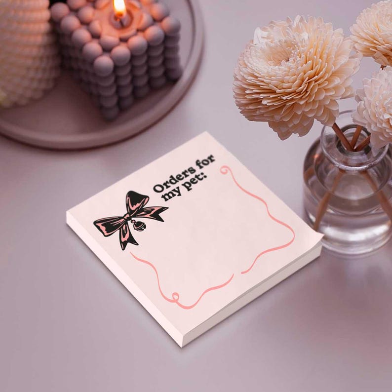 Peut inclure: Un bloc-notes carr&eacute; rose clair avec le texte "Orders for my pet:" et une illustration d'un n&oelig;ud noir. Le bloc-notes a une bordure rose ondul&eacute;e. Des fleurs d&eacute;coratives et une bougie sont en arri&egrave;re-plan.