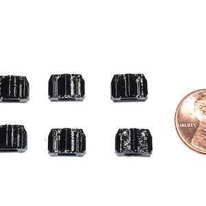May include: Six small, black, rectangular objects are arranged on a white surface, next to a penny. The objects have a ridged texture. The penny shows a profile of a person and the word "Liberty".