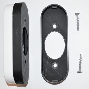 May include: A white and black rectangular device with an oval opening, next to a black oval frame and two grey screws. The device has a rounded edge and appears to be made of plastic. The frame has two holes for screws.