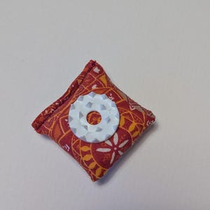 Könnte beinhalten: Ein kleines, quadratisches Nadelkissen aus Stoff mit einem rot-orangen geometrischen Blumenmuster. In der Mitte des Kissens befindet sich ein weißes, kreisförmiges Kunststoff-Nähwerkzeug mit gezacktem Rand und einem Loch in der Mitte.