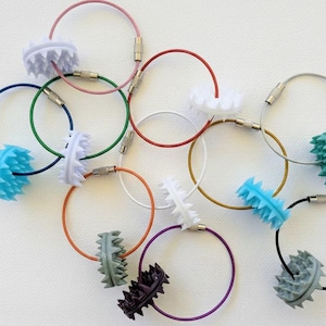 Könnte beinhalten: Eine Sammlung von runden Drahtschlüsselringen in verschiedenen Farben wie Blau, Grün, Orange, Rot, Weiß, Lila und Schwarz, wobei jeder Ring mit einer kleinen, stacheligen, runden Silikonperle in passender oder kontrastierender Farbe versehen ist.