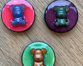 May include: Three round phone grips with a glittery resin finish. Each grip features a small, detailed bear figurine in a different colour: blue, green, and brown. The grips are set against a wood background.