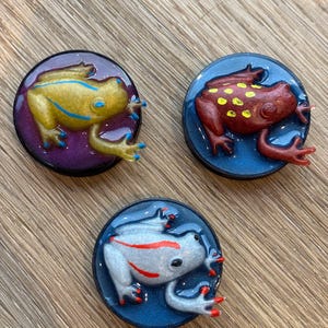 May include: Three resin frog figurines, each with a different colour scheme. The first frog is yellow with blue stripes, the second is red with yellow spots, and the third is white with red stripes. Each frog is sitting on a round, coloured base.