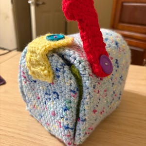 Könnte beinhalten: Eine handgefertigte, gehäkelte Briefkasten aus hellblauem, gesprenkeltem Garn. Der Briefkasten hat eine rote Fahne, eine gelbe Klappe und bunte Knöpfe. Das Gesamtdesign ist skurril und dekorativ.