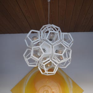 Könnte beinhalten: Eine weiße geometrische Pendelleuchte mit mehreren Glühbirnen. Die Leuchte besteht aus einer Reihe von miteinander verbundenen sechseckigen Formen.