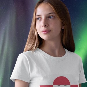 Peut inclure: T-shirt blanc avec un motif graphique d'un cercle rouge sur une forme horizontale rouge et blanche avec un visage souriant. Le t-shirt est port&eacute; par une personne aux longs cheveux bruns, sur fond de ciel &eacute;toil&eacute; aux teintes vertes et violettes.