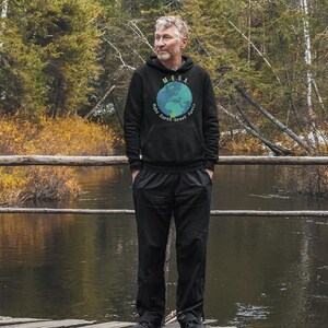 Peut inclure: Une personne portant un sweat-shirt &agrave; capuche noir avec un globe terrestre bleu et vert et le texte "M.E.G.A. Make Earth Great Again". La personne est debout sur un pont en bois au-dessus d'une rivi&egrave;re.