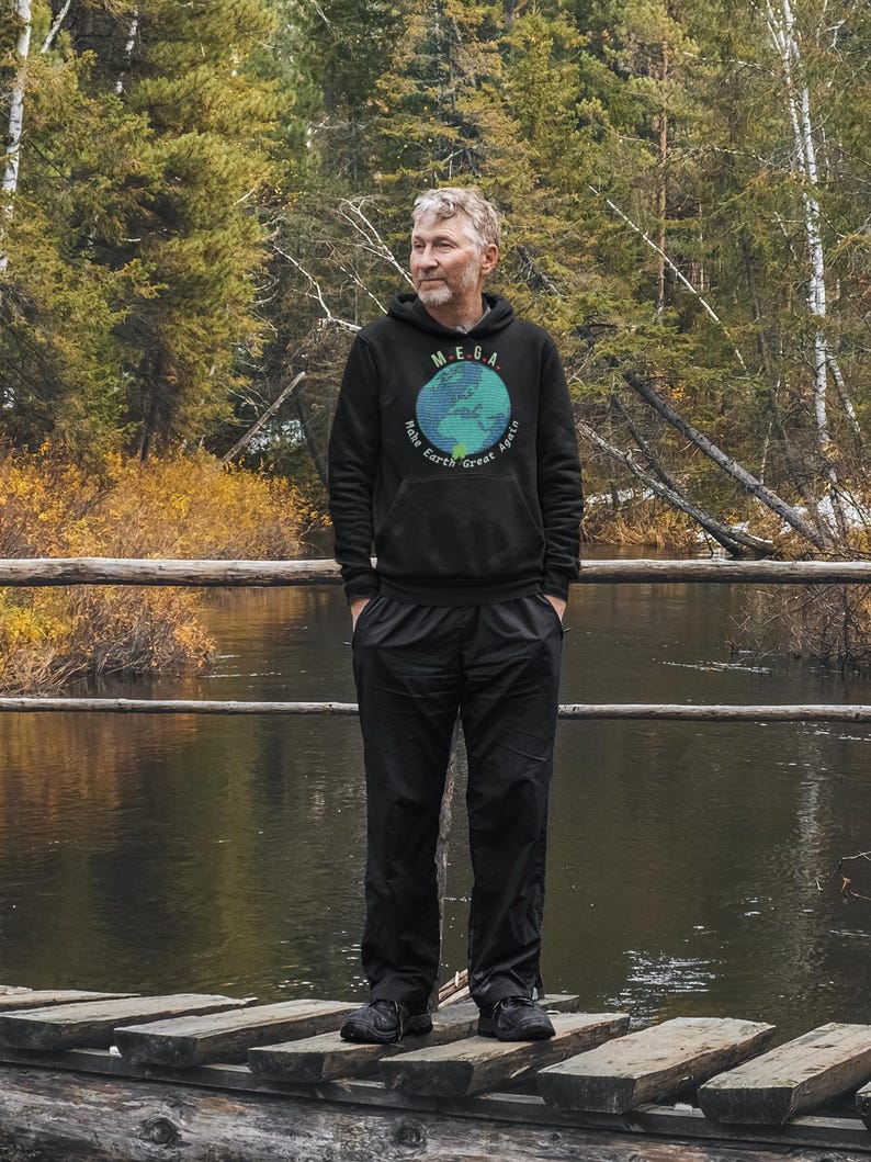 Peut inclure: Une personne portant un sweat-shirt &agrave; capuche noir avec un globe terrestre bleu et vert et le texte "M.E.G.A. Make Earth Great Again". La personne est debout sur un pont en bois au-dessus d'une rivi&egrave;re.