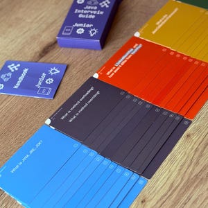 Puede incluir: Un conjunto de tarjetas y una peque&ntilde;a caja sobre una superficie de madera. La caja es morada y lleva la etiqueta "Java Interview Guide Junior". Las tarjetas son azules, rojas, amarillas y negras, con texto como "What is JVM, JRE, JDK?"