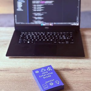 Puede incluir: Un portátil con código en la pantalla está sobre una superficie de madera. Delante hay una caja morada con el texto "Java Interview Guide Junior" y varios iconos. El portátil tiene un teclado negro.