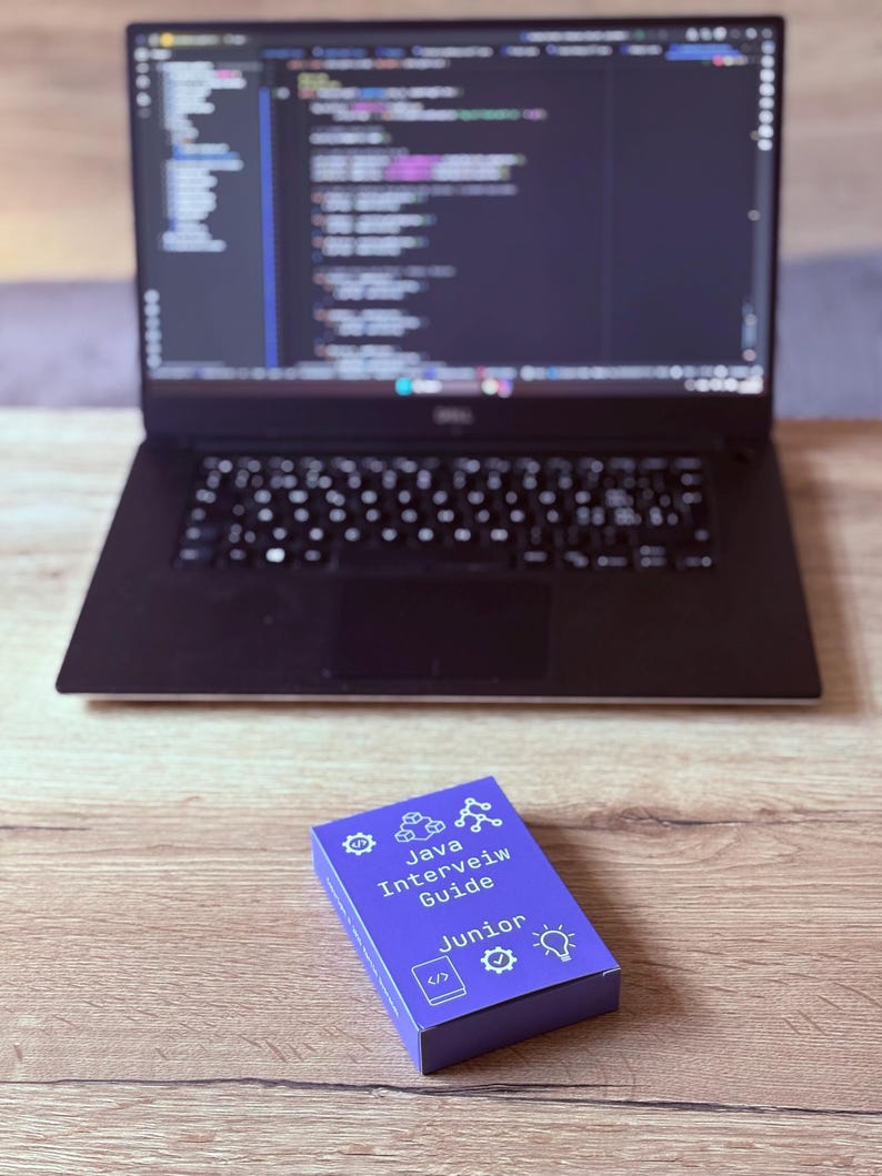 Puede incluir: Un port&aacute;til con c&oacute;digo en la pantalla est&aacute; sobre una superficie de madera. Delante hay una caja morada con el texto "Java Interview Guide Junior" y varios iconos. El port&aacute;til tiene un teclado negro.