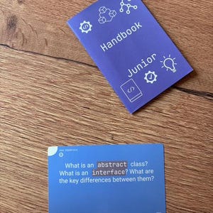 Puede incluir: Un folleto azul "Handbook Junior" y una tarjeta azul con el texto "What is an abstract class? What is an interface? What are the key differences between them?" sobre una superficie de madera.