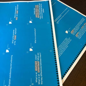 Puede incluir: Varios documentos educativos azules con texto blanco est&aacute;n apilados sobre una superficie marr&oacute;n oscuro. Los documentos est&aacute;n encuadernados en espiral y contienen preguntas sobre conceptos de programaci&oacute;n.