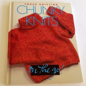 Könnte beinhalten: Ein Buch mit dem Titel "Vogue Knitting Chunky Knits" mit einem rot-orangen Strickpullover auf dem Cover. Die Worte "Chunky Knits" sind in großen blauen Buchstaben geschrieben, und die Worte "on the go!" sind in blauer Schrift. Das Buch ist auf weißem Hintergrund.