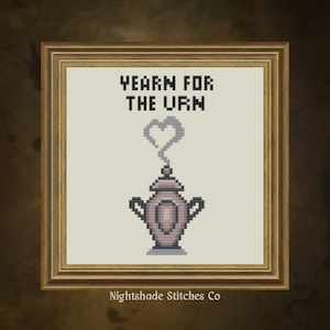Op de afbeelding: Kruissteekkunst in een gouden lijst. Het kunstwerk toont een roze urn met stoom in de vorm van een hart en de tekst "YEARN FOR THE URN". De naam van de kunstenaar, "Nightshade Stitches Co", staat onderaan.