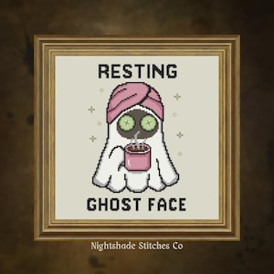 May include: A framed cross-stitch pattern featuring a white ghost wearing a pink towel wrap on its head and cucumber slices over its eyes. The ghost holds a pink mug with steam rising. The text reads RESTING GHOST FACE in black block letters.