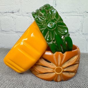 Puede incluir: Una variedad de joyas vintage de baquelita. La imagen presenta un brazalete amarillo brillante, un recipiente tallado en forma de flor naranja y un peine para el cabello floral verde. Las piezas están dispuestas sobre una superficie gris estampada.