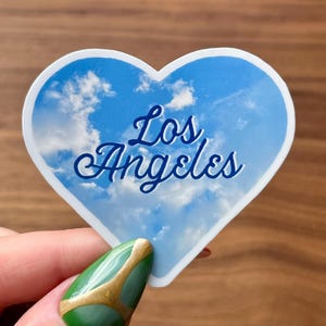 May include: A heart-shaped sticker featuring a blue sky with fluffy white clouds. The cursive text "Los Angeles" is written in blue across the center of the sticker. The sticker is held by a hand with a green and gold nail design.