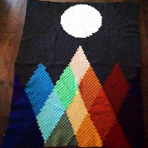 May include: A handmade crochet blanket featuring a night sky design with a large white moon and colorful mountain range. The mountains are in shades of blue, green, yellow, orange, and red. The blanket is approximately 60 inches by 48 inches.