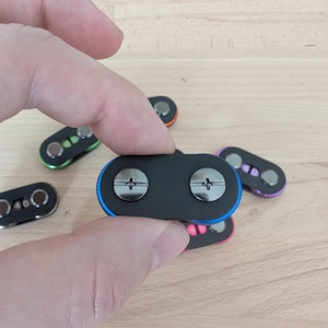May include: A hand holding a black magnetic clasp with two silver screws. The clasp has a blue border. Other clasps with different colored borders, including green, orange, pink, and purple, are visible in the background.