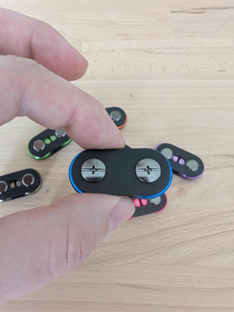May include: A hand holding a black magnetic clasp with two silver screws. The clasp has a blue border. Other clasps with different colored borders, including green, orange, pink, and purple, are visible in the background.