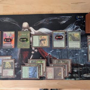 May include: A Magic: The Gathering playmat with a dark background featuring a fantasy illustration. Several game cards are laid out, along with a deck of cards and card holders. The cards have various illustrations and text.