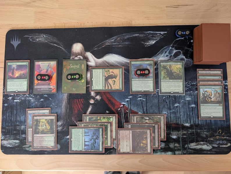 May include: A Magic: The Gathering playmat with a dark background featuring a fantasy illustration. Several game cards are laid out, along with a deck of cards and card holders. The cards have various illustrations and text.