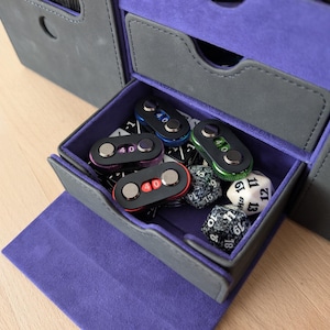 May include: A gray and purple gaming accessory storage box with a drawer open, revealing dice and counters. The drawer contains several black counters with the number "40" and dice.