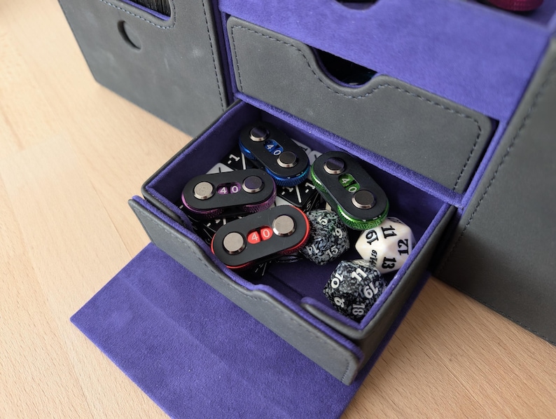 May include: A gray and purple gaming accessory storage box with a drawer open, revealing dice and counters. The drawer contains several black counters with the number "40" and dice.